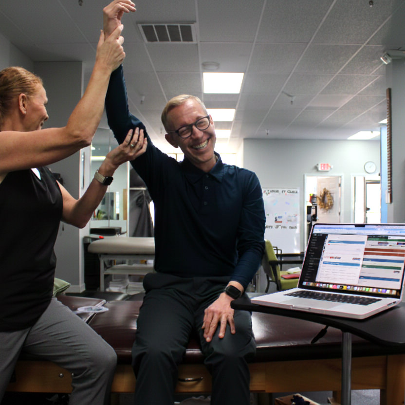 A person sits on an exam table smiling while a healthcare professional raises their arm; a laptop with open tabs for the AI Scribe landing page sits beside them.